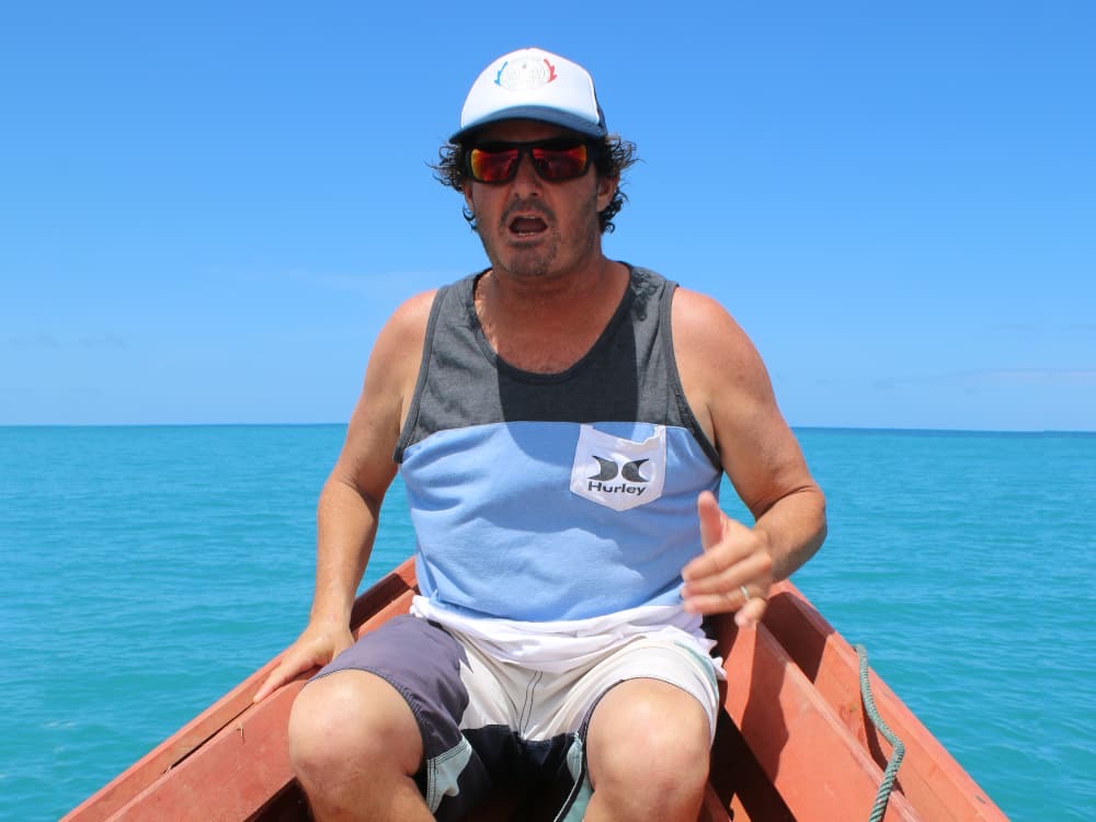 Dave Renaud sitting in a wooden boat on flat turquoise water, mid-sentence, throwing a shaka