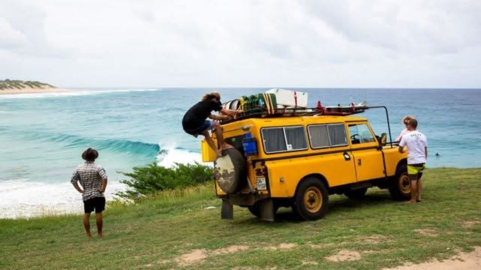 A yellow Land Rover loaded with boards on a grassy headland above a pointbreak